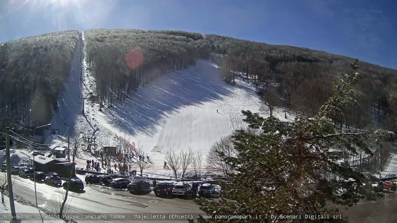 A snow-covered ski slope on a sunny day, with a ski lift and several parked cars visible near the base.