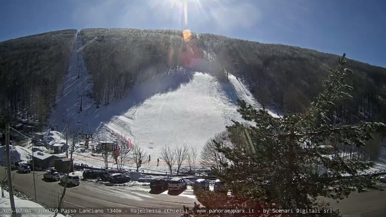 A sunny winter day at a ski slope shows a snow-covered hill with a ski lift and several people skiing, viewed from a parking lot below.