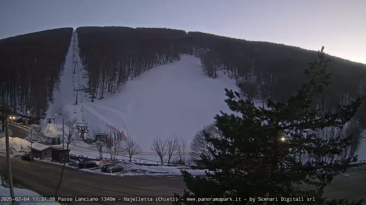 A snow-covered ski slope with a chairlift ascends a wooded hill under a twilight sky.