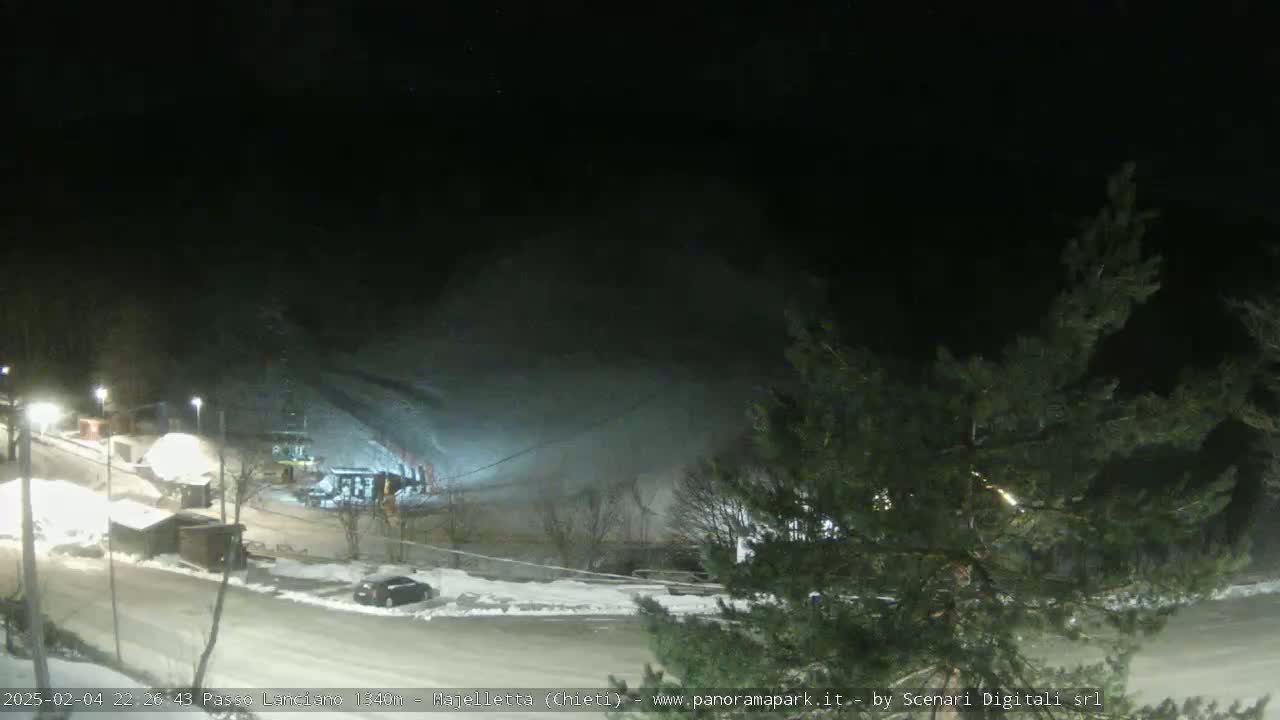 A nighttime view of a snow-covered parking lot and ski slope, illuminated by artificial lights, with a hazy or foggy atmosphere.