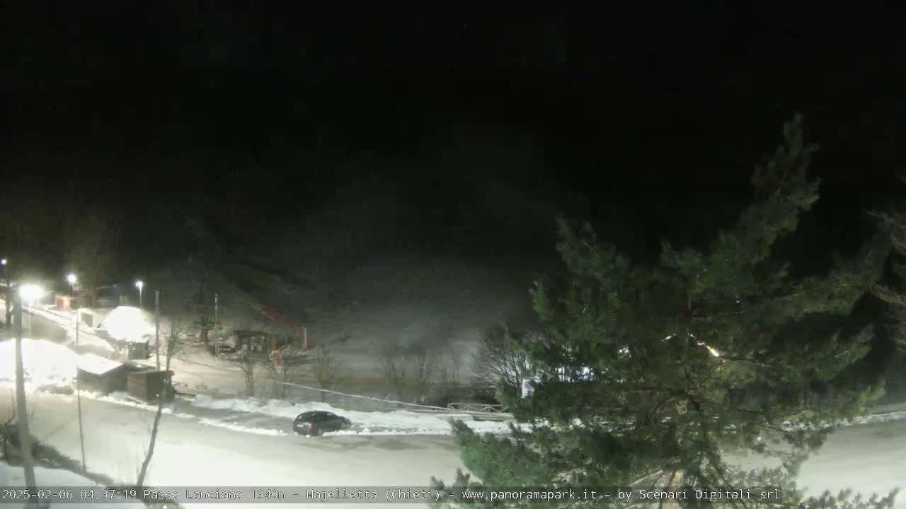 A nighttime, snow-covered scene shows a parking lot with a few cars, some small buildings, and a sparsely lit area with some trees.