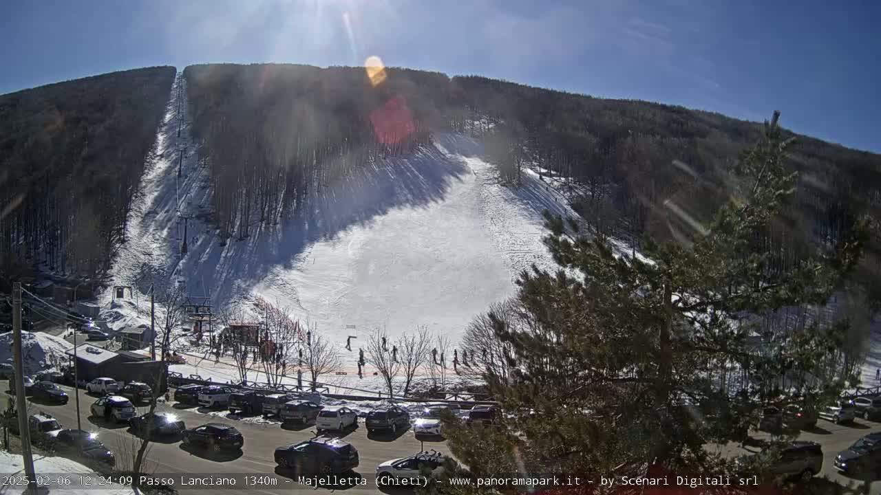 A sunny day at a snow-covered ski slope with a chairlift, several skiers, and a parking lot full of cars.