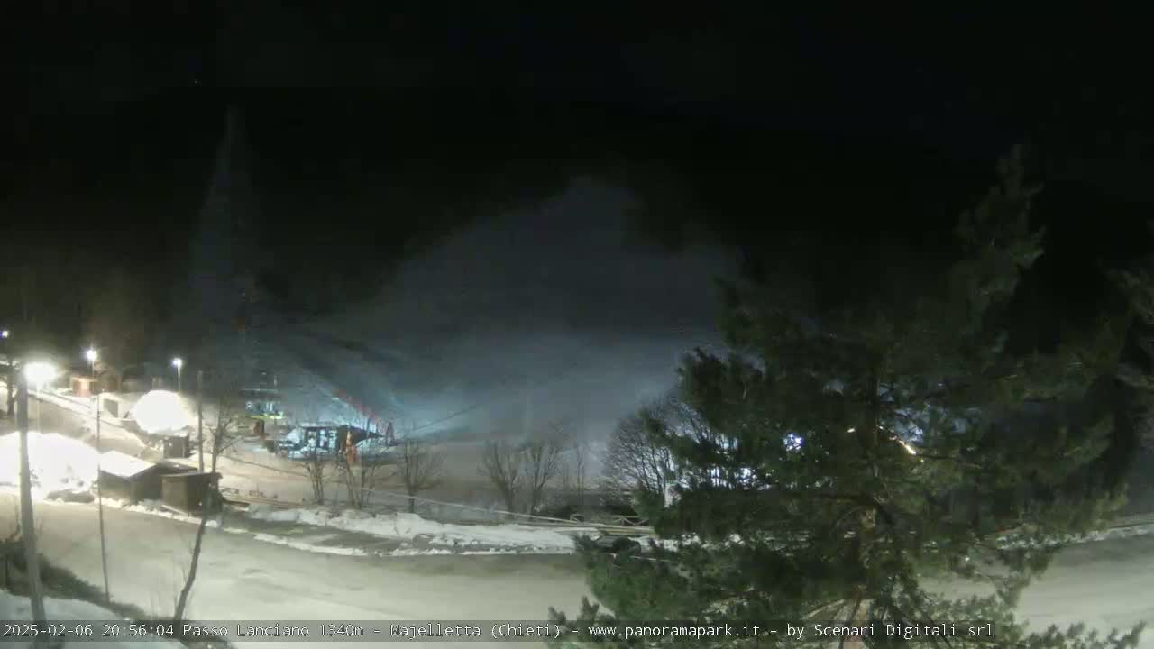 A nighttime view of a snow-covered ski slope with snow blowing across it, illuminated by artificial lights, and surrounded by dark trees.