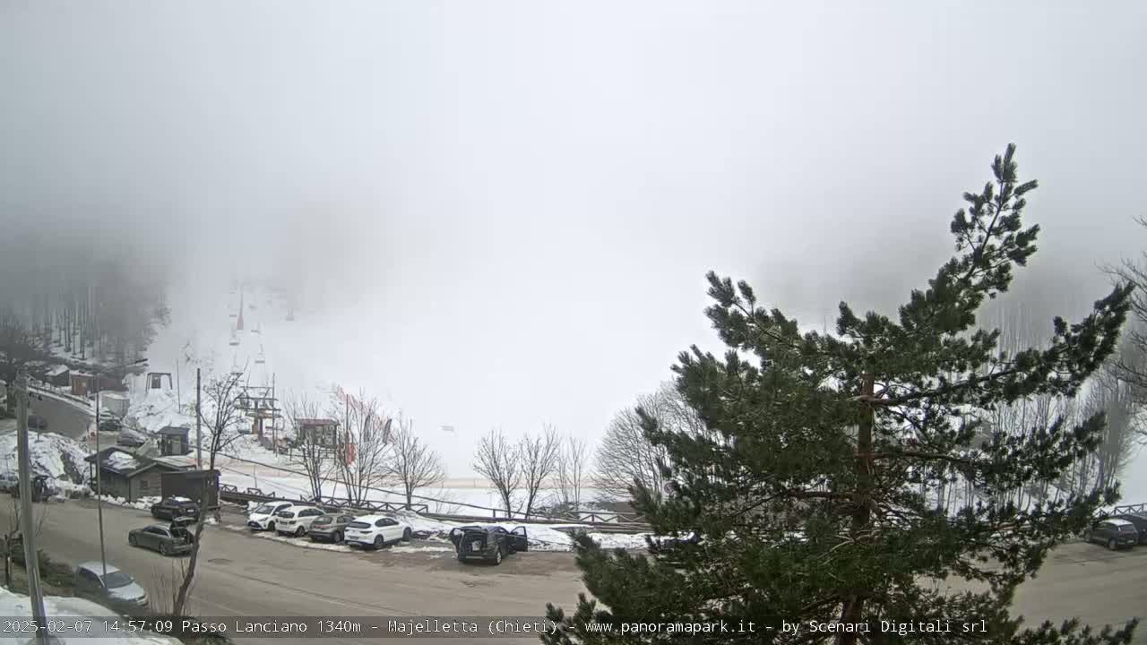 A foggy, snowy mountain scene shows a parking lot with several cars near a ski lift and snow-covered trees.