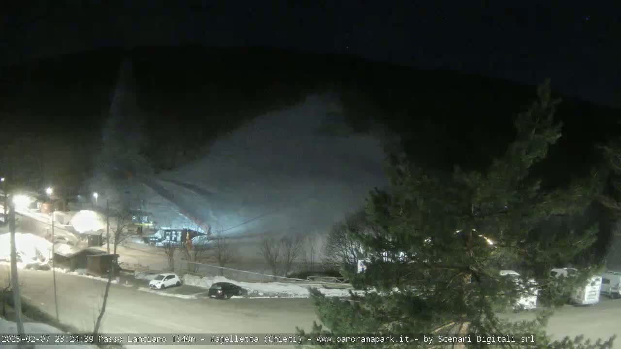 A nighttime snowmaking operation illuminates a ski slope and parking lot, with snow-covered ground and dark evergreen trees visible in the foreground.