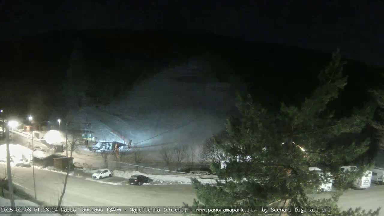 A nighttime, snow-covered parking lot and roadway with several vehicles, a ski lift, and some buildings are visible under a dark sky.