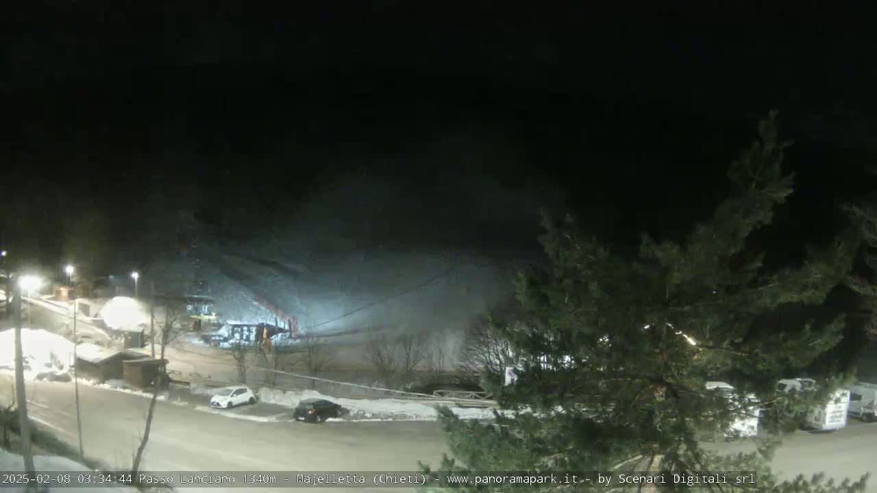 A nighttime scene of a snow-covered hill with a parking lot containing several cars and RVs, illuminated by artificial lights.
