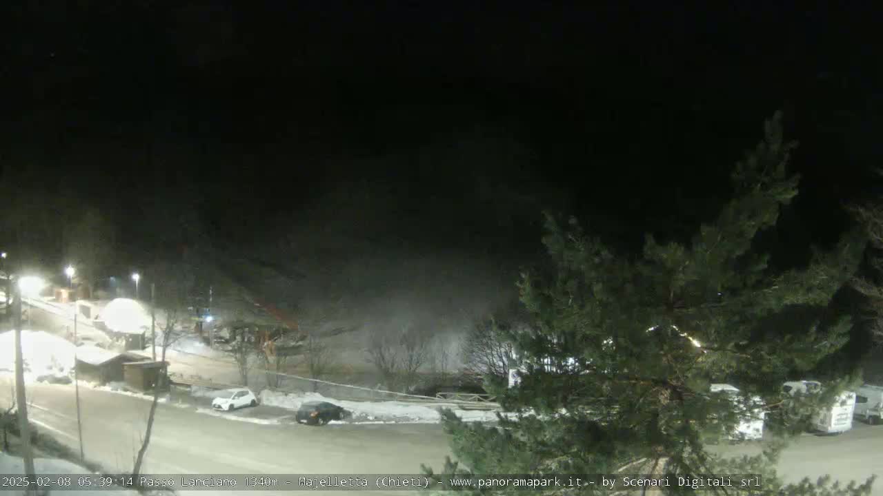 A nighttime, snow-covered parking lot and roadway are dimly lit, with a few vehicles present and some buildings in the background partially obscured by fog or mist.