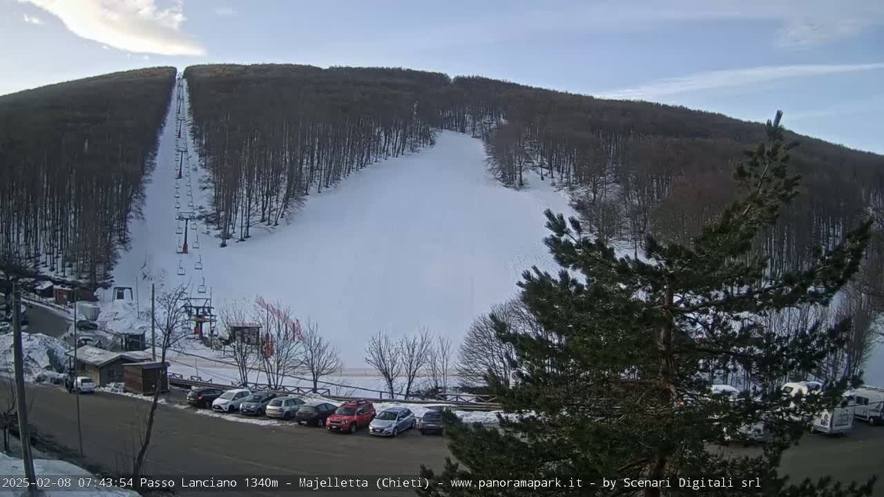 A snow-covered ski slope on a mountainside with a chairlift and a parking lot of cars below under a mostly clear sky.