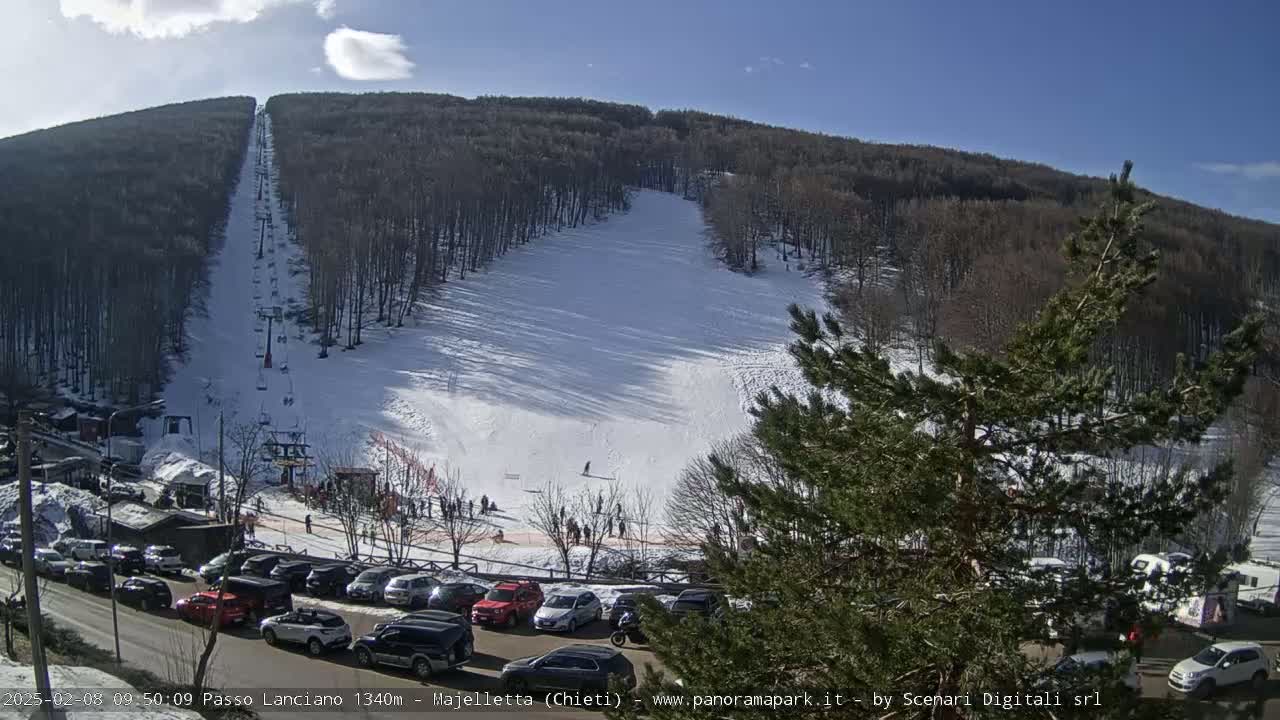 A snow-covered ski slope with a chairlift and a parking lot full of cars is visible on a sunny day.