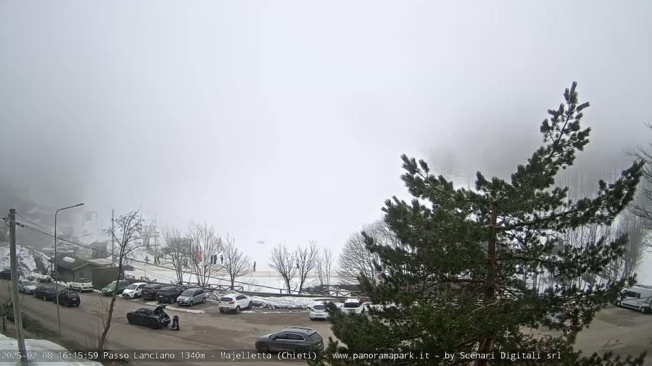 A foggy mountain parking lot with cars and a few people visible, surrounded by snow-covered trees and a building.
