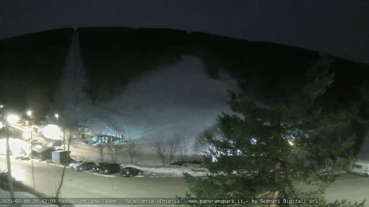 A nighttime view of a snow-covered ski slope with snow being blown from a snow-making machine, illuminated by artificial lights, with cars parked in a lot below.
