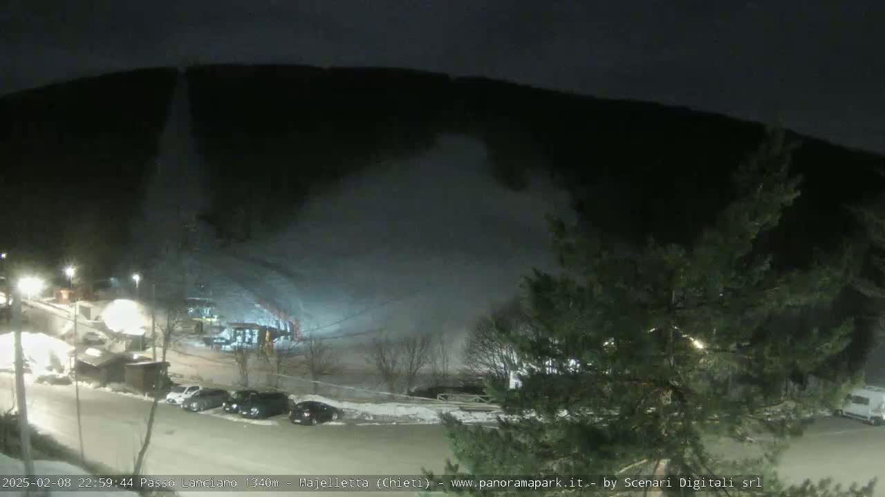 A nighttime view of a snow-covered hill with a snowmaking machine operating, a parking lot with cars, and some buildings, under a dark sky.