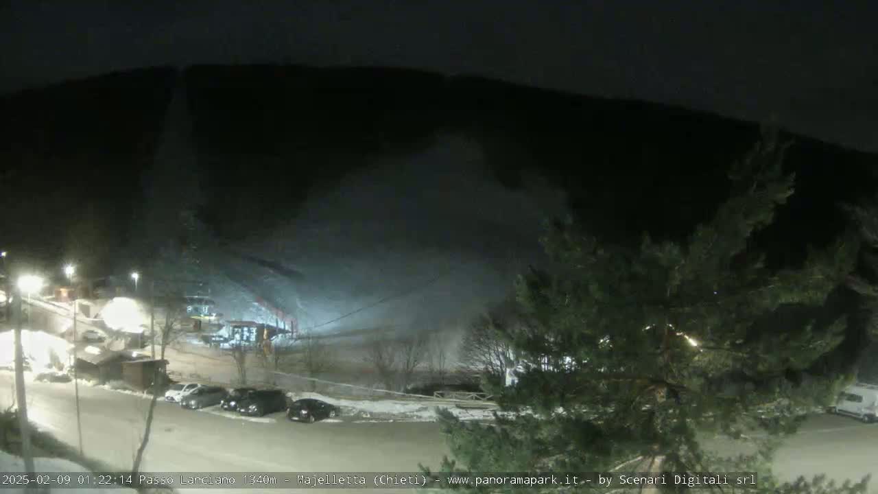 A nighttime, snow-covered parking lot with cars and buildings sits below a snow-covered hill and dark, wooded mountainside.