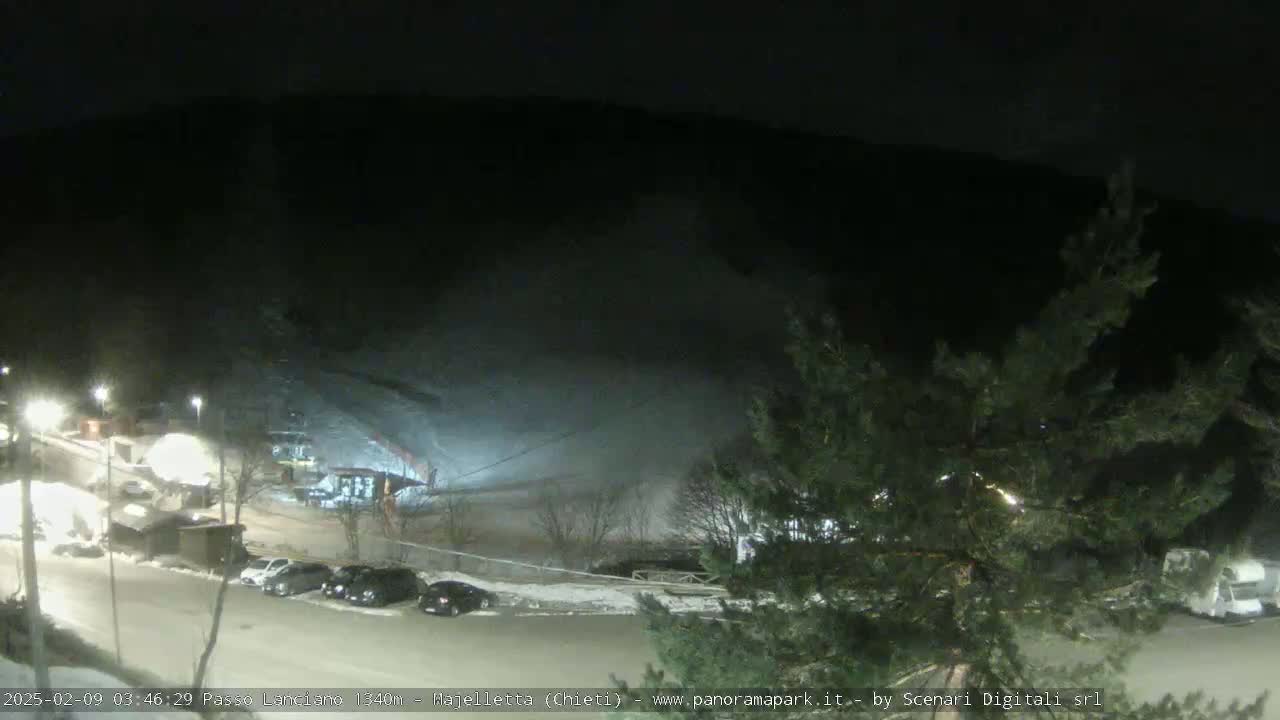 A nighttime, snow-covered parking lot with cars and buildings is visible below a dark, snow-covered hillside.