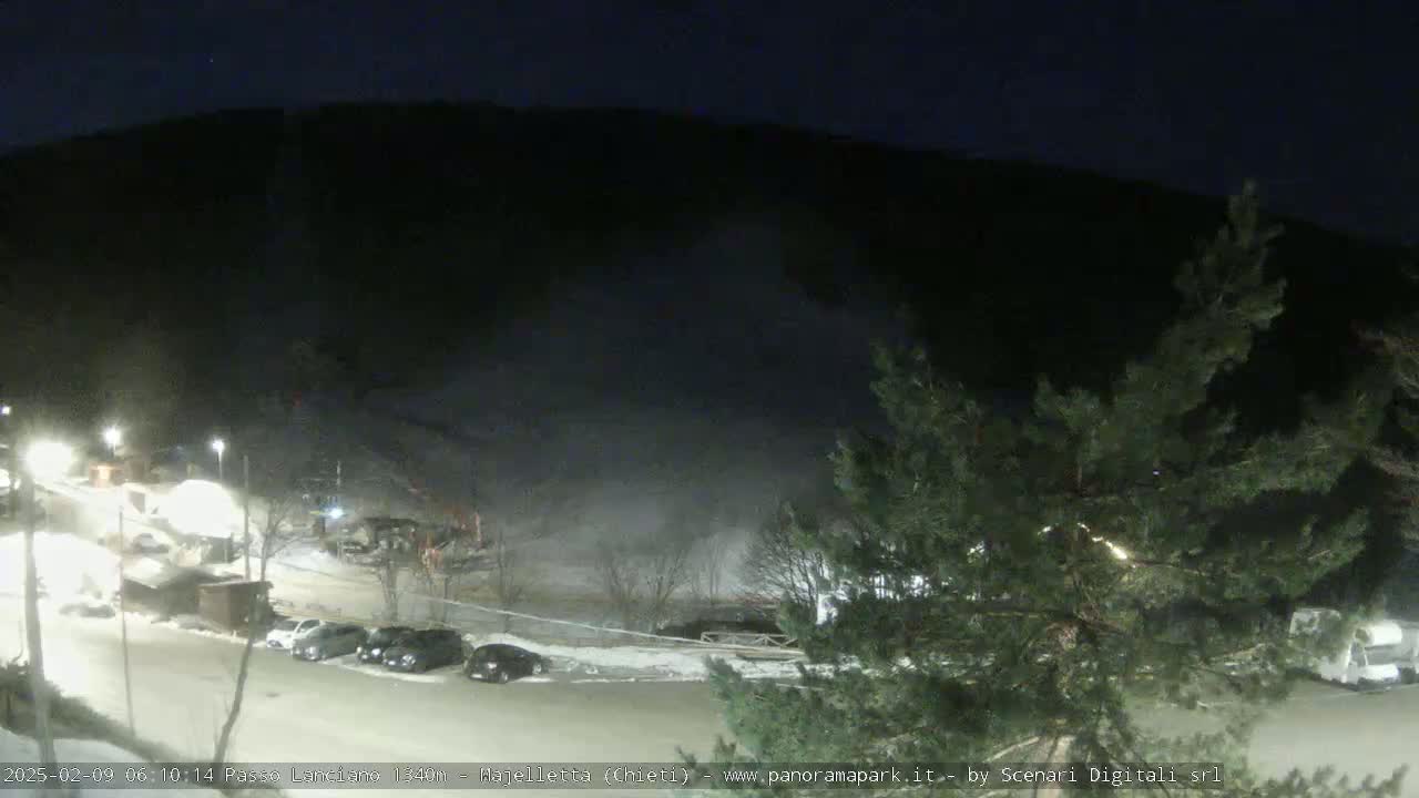 A nighttime, snow-covered parking lot with several cars and buildings is illuminated by lights against a dark mountain background.