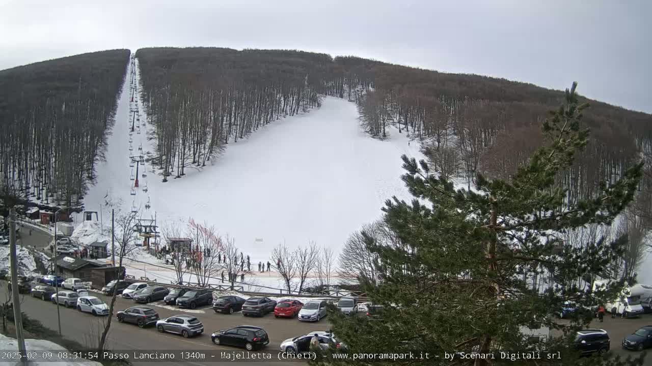 A snow-covered ski slope with a chairlift and several parked cars is visible, surrounded by a wooded hillside under an overcast sky.