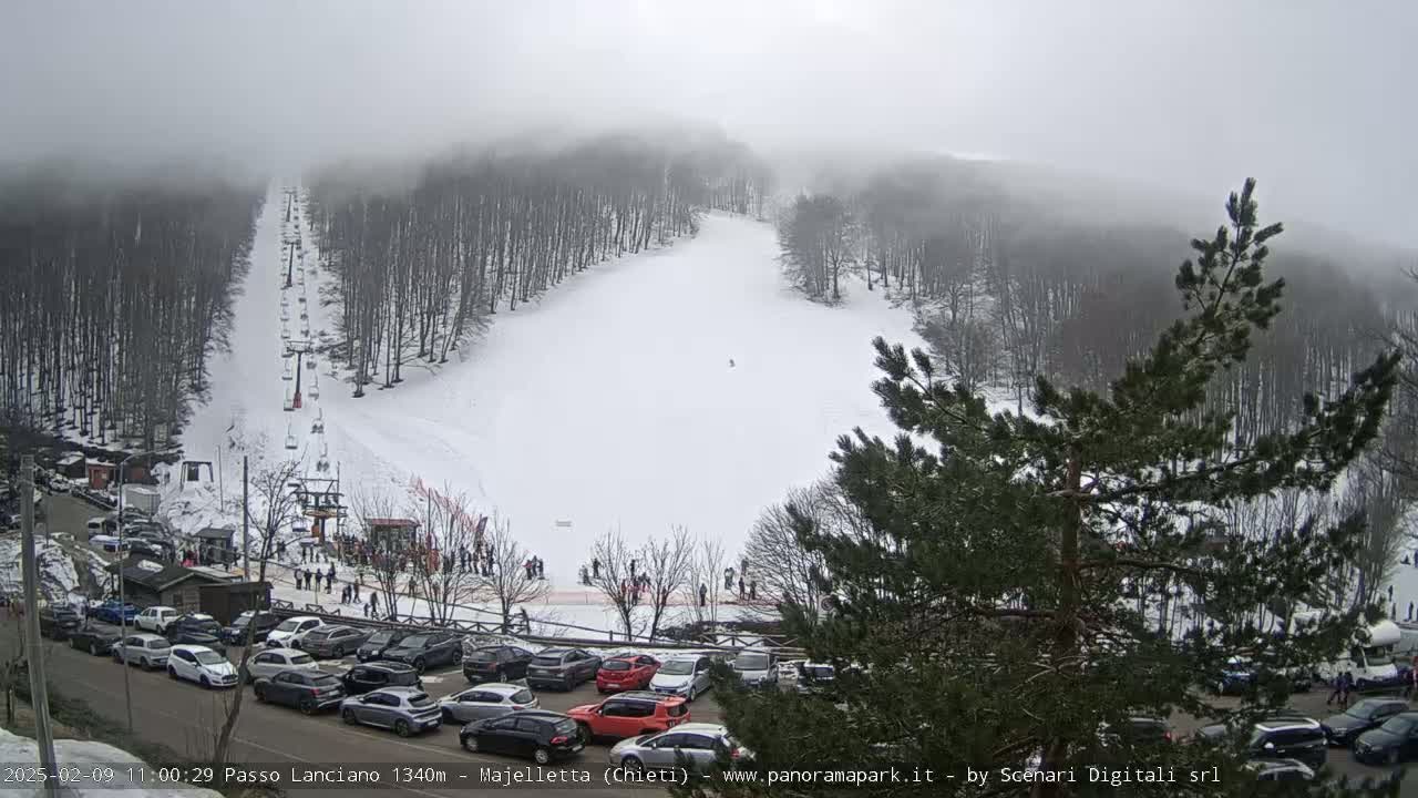 A snowy ski slope with a chairlift and a parking lot full of cars is visible under a foggy sky.