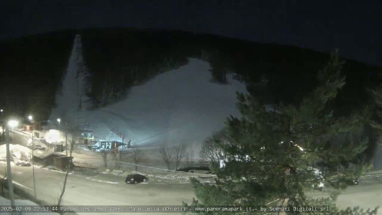 A nighttime view of a snow-covered ski slope, illuminated by artificial lights, with a few cars parked in a lot below.