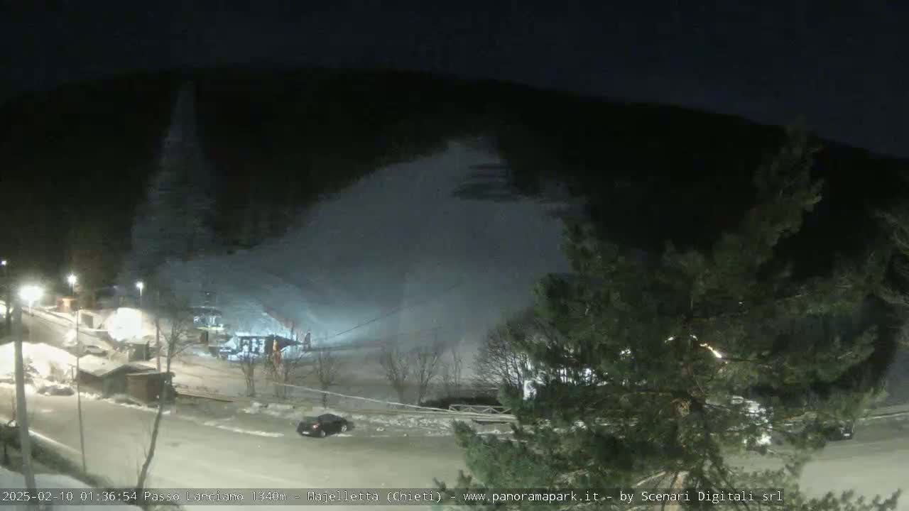 A nighttime view of a snow-covered ski slope with a few buildings and a car in a parking lot below, under a dark sky.