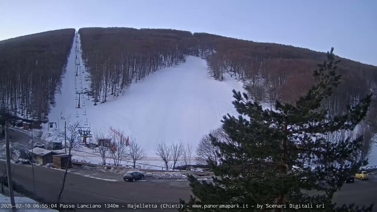 A ski slope on a snow-covered hillside with a chairlift and a few cars in a parking lot under a clear, twilight sky.