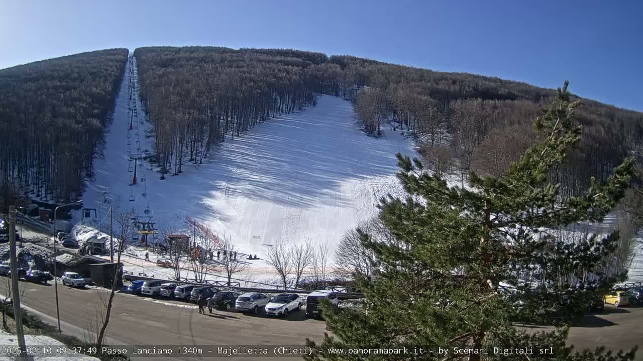 A ski slope on a sunny day, covered in snow, with a chairlift running up its length, and cars parked in a lot at the bottom.