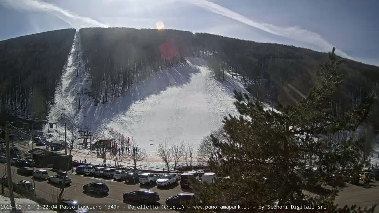 A sunny day reveals a snow-covered ski slope with a ski lift, a parking lot full of cars, and a wooded hillside.
