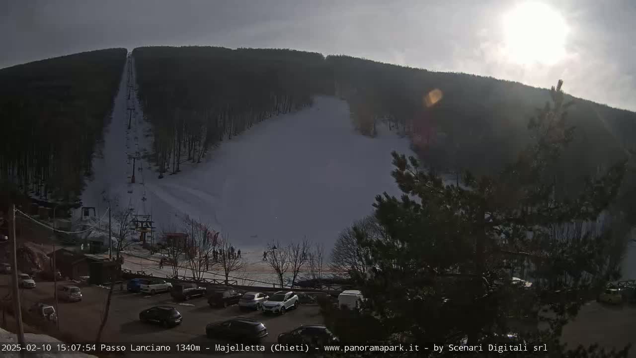 A sunny day at a snow-covered ski slope with a ski lift visible, and a parking lot with several cars below.