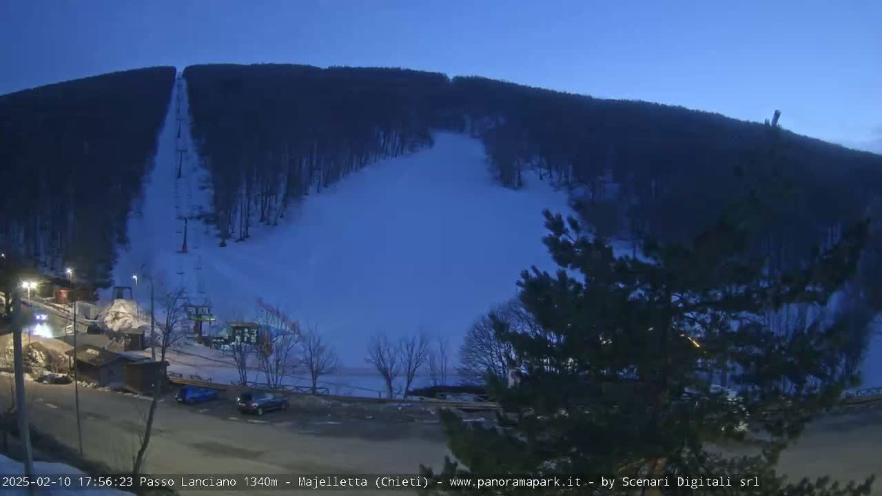 A ski slope on a snow-covered hill at dusk, with a chairlift visible and a few cars parked in the lot below.