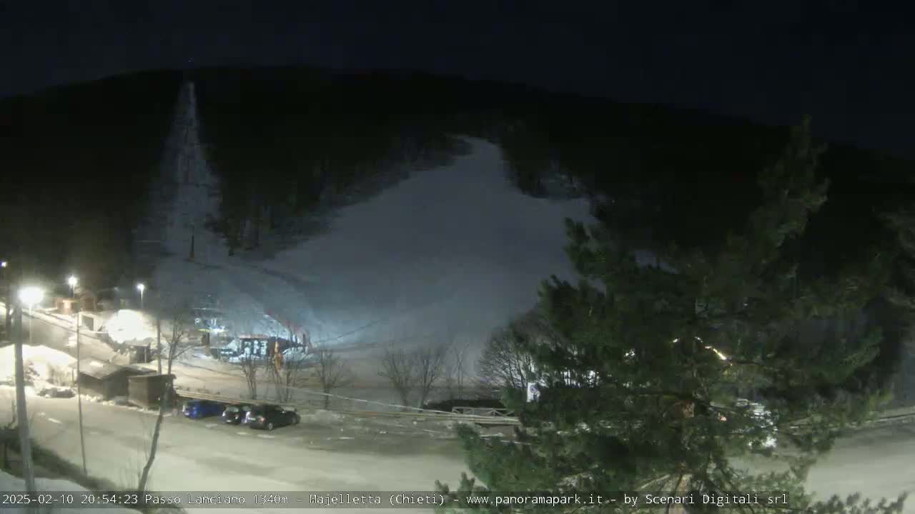 A nighttime view of a snow-covered hillside with a few cars parked in a lot below, illuminated by artificial lights.