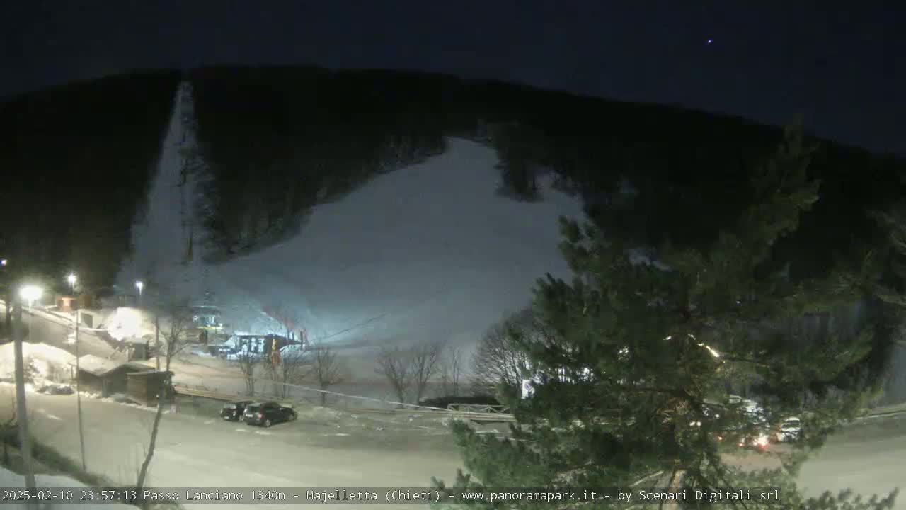 A snow-covered ski slope at night, illuminated by artificial lights, with several vehicles parked in a lot at the bottom of the hill.