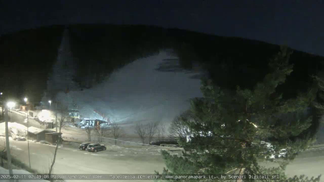A nighttime, snow-covered ski slope is visible, with a parking lot and some buildings illuminated in the foreground.
