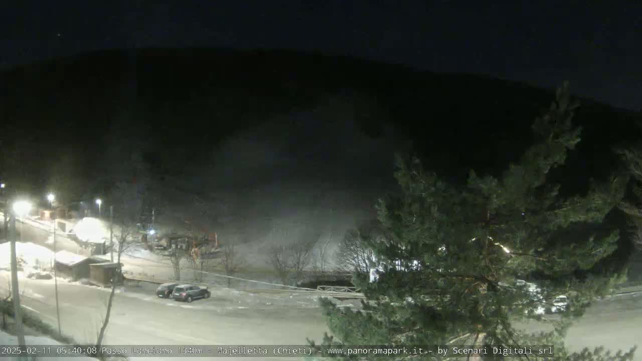 A nighttime, snow-covered parking lot and road are illuminated, with a dark mountainside rising in the background under a clear night sky.