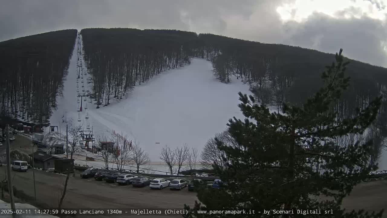 A snow-covered ski slope with a chairlift and a parking lot of cars underneath an overcast sky.