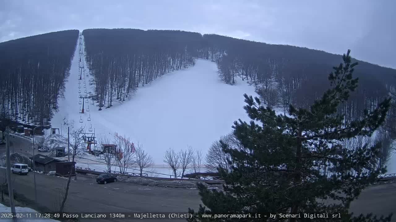 A snow-covered ski slope with a chairlift extends up a mountainside under an overcast sky.
