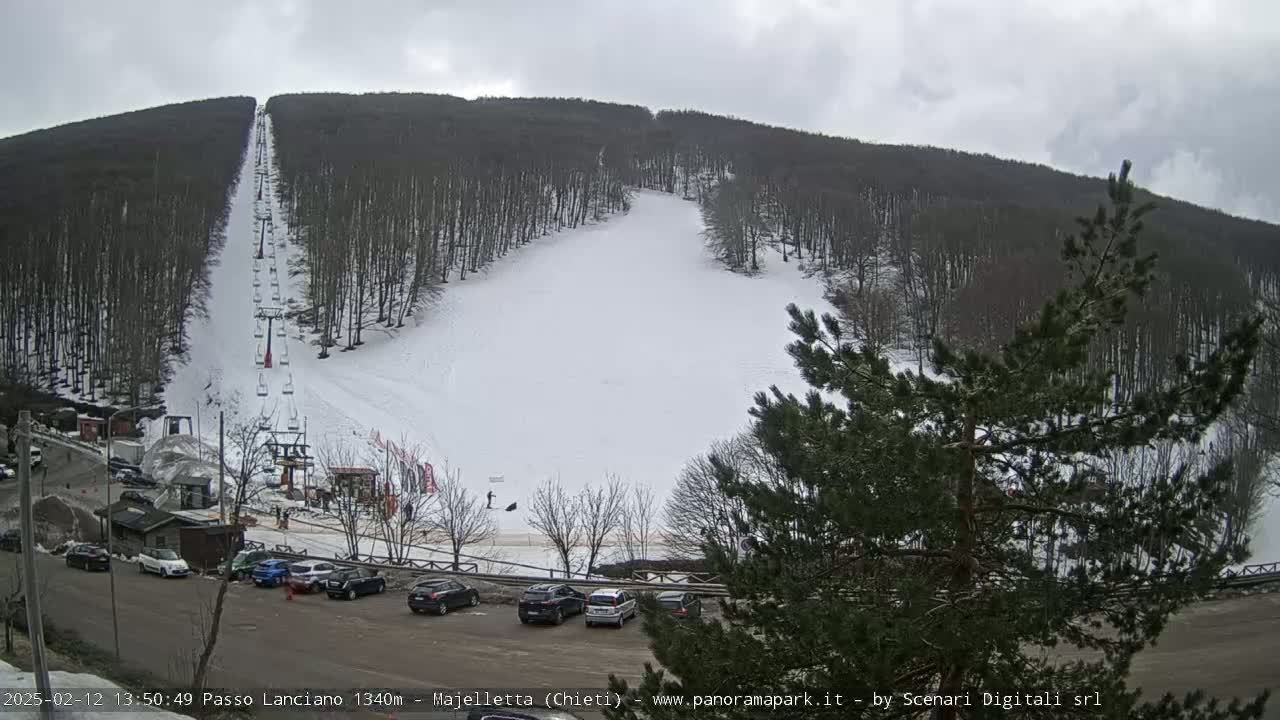 A snow-covered ski slope with a chairlift is visible on a cloudy day, with cars parked in a lot below.