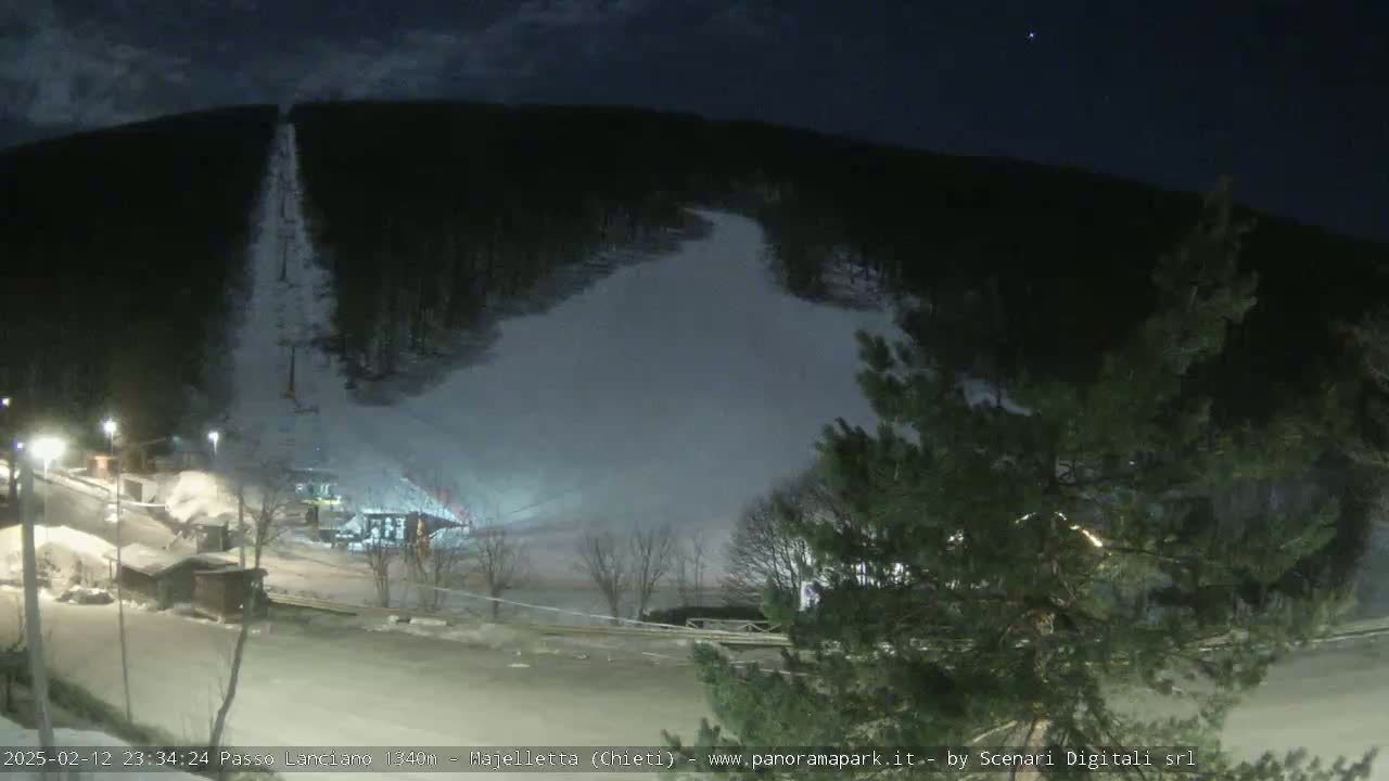 A nighttime view of a snow-covered ski slope with a chairlift and some buildings at the base, under a mostly clear sky.