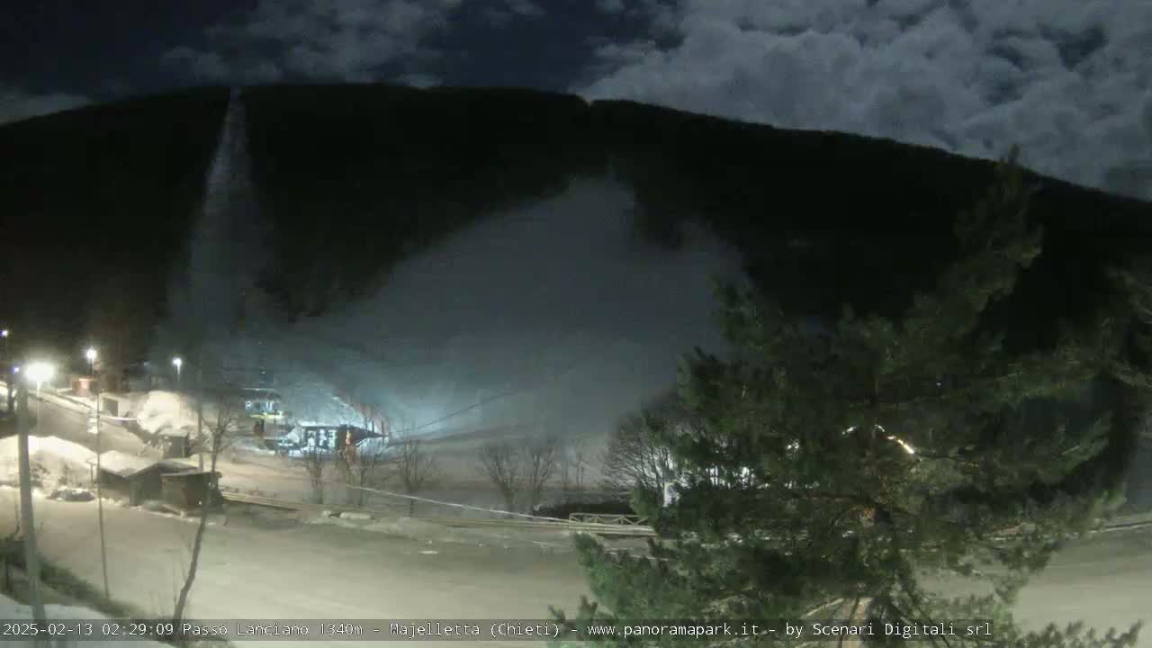 A nighttime view of a snow-covered ski slope with a snow-making machine operating under a partly cloudy sky.
