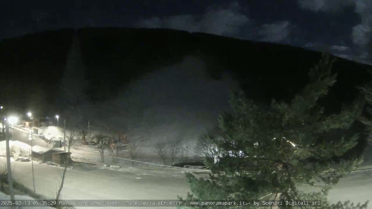 A nighttime, snow-covered landscape shows a mountainside under a partly cloudy sky, with a few lights illuminating a small building complex and surrounding area.