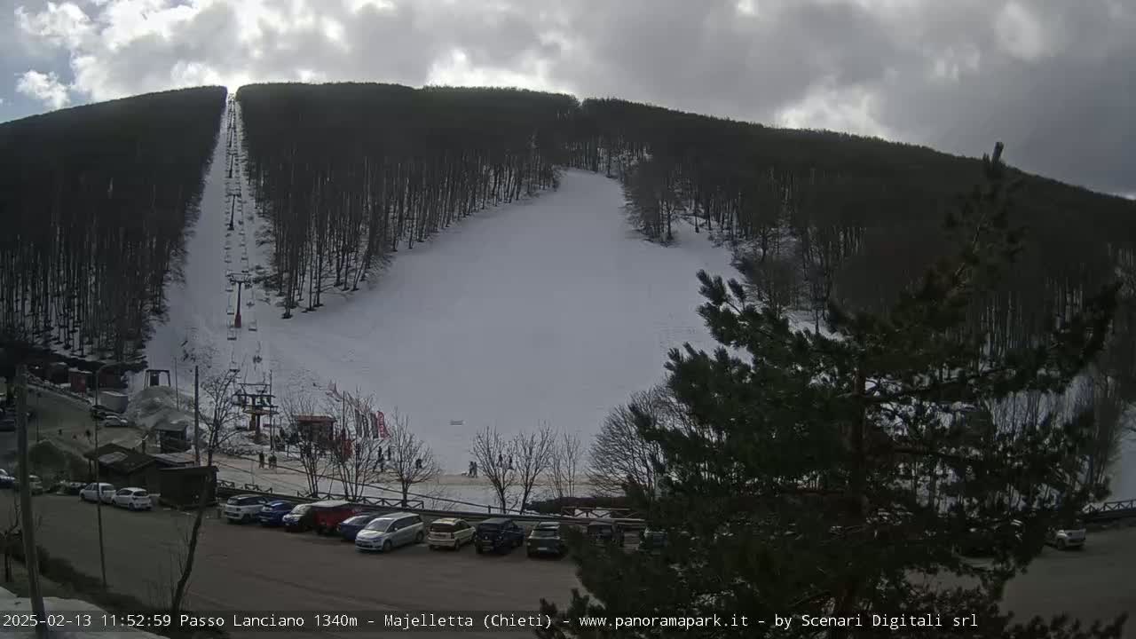 A snow-covered ski slope with a chairlift ascends a wooded hillside under a partly cloudy sky.