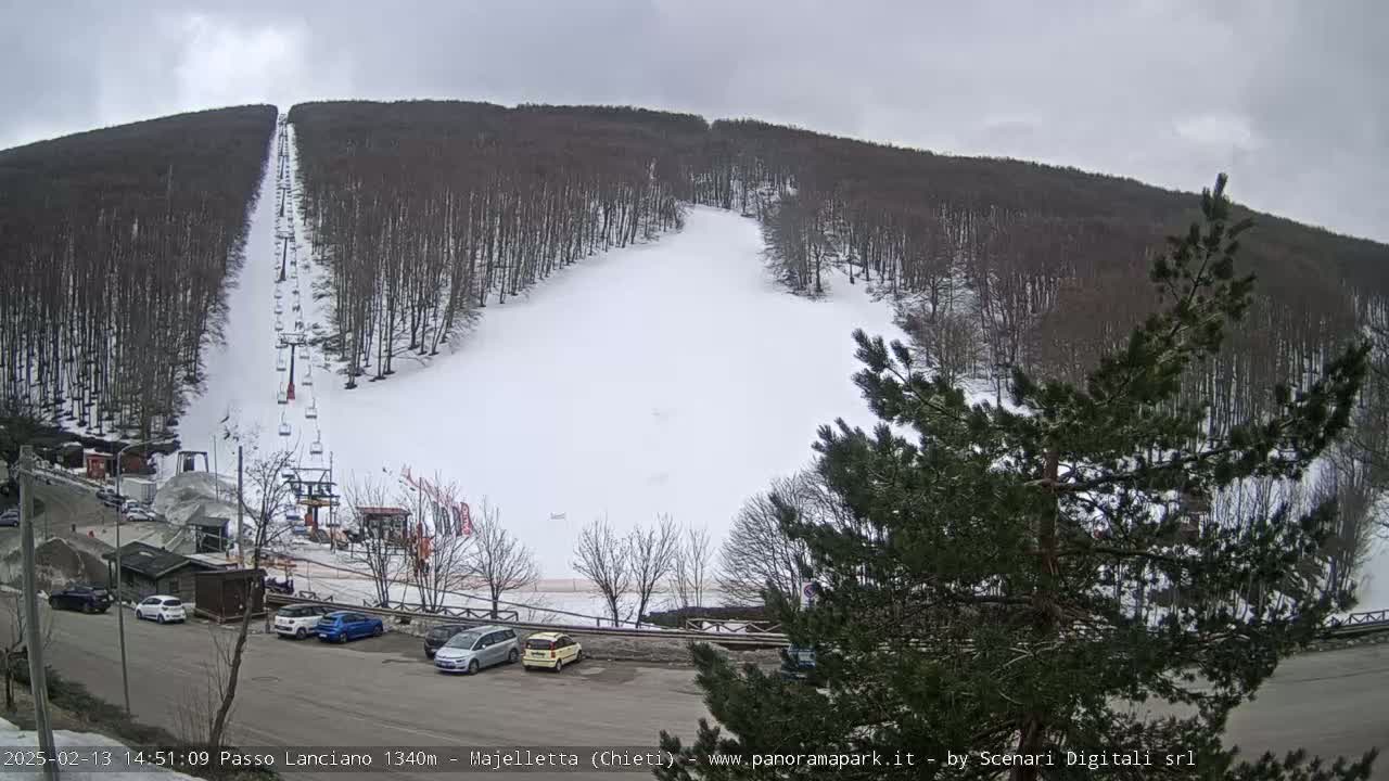 A snow-covered ski slope with a chairlift is visible between two forested hills on an overcast day.
