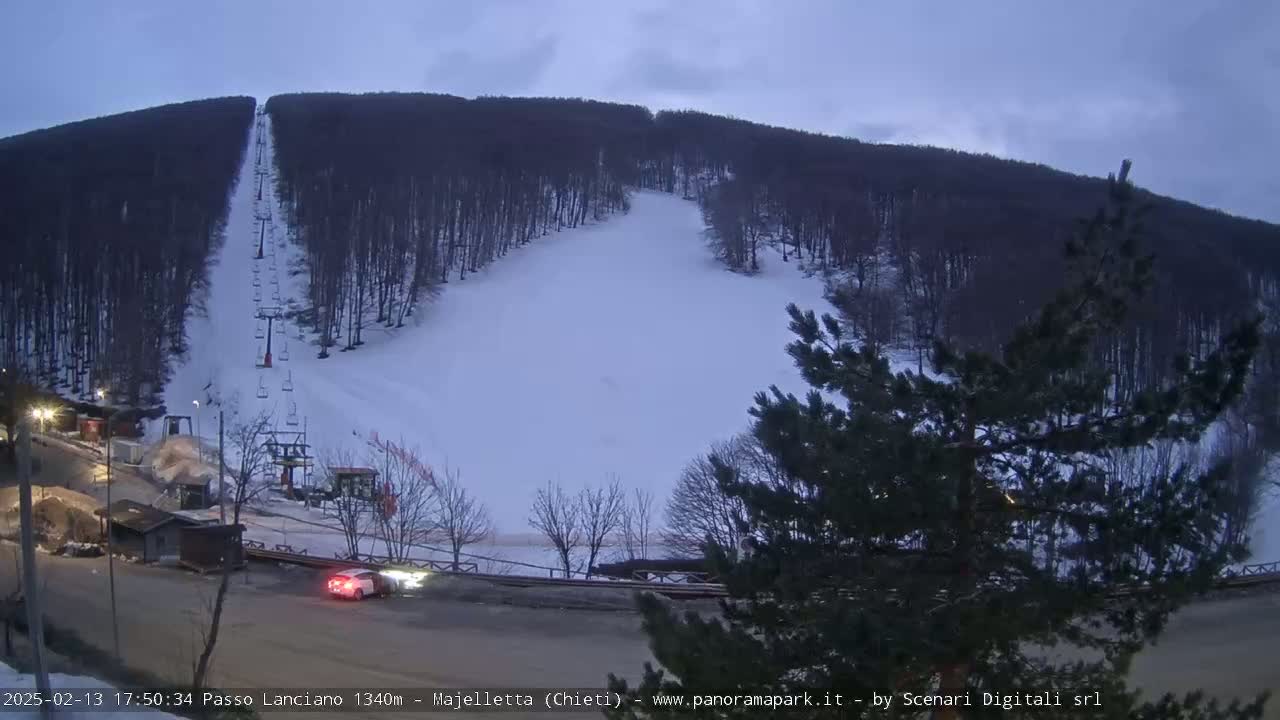 A snow-covered ski slope with a chairlift and a few cars at the base under a twilight sky.