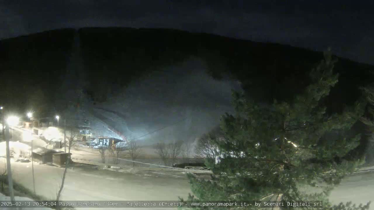 A nighttime, snowy scene shows a hill with lights and structures at its base, illuminated by artificial light, with snow blowing faintly.