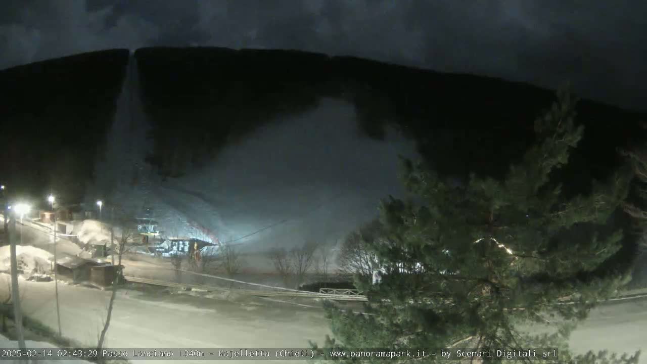 A nighttime scene of snow being blown from a hillside onto a roadway below, with some trees and buildings visible in the foreground under a dark sky.
