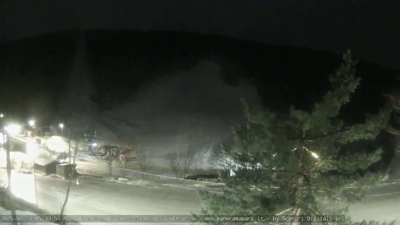 A nighttime, snowy scene shows a parking lot and a hill shrouded in blowing snow.