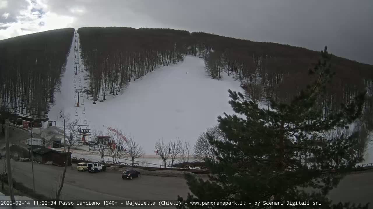 A snow-covered ski slope with a chairlift runs through a wooded area under an overcast sky.