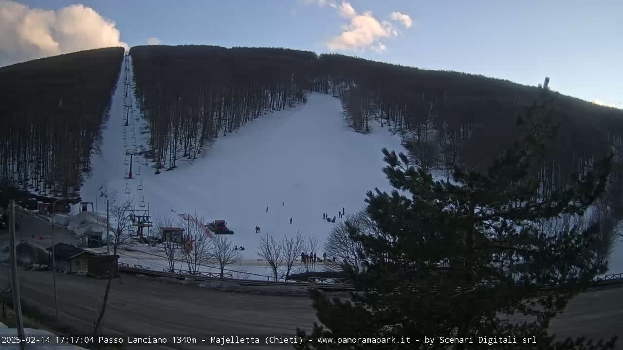 A snow-covered ski slope with a chairlift and several skiers visible under a partly cloudy sky.
