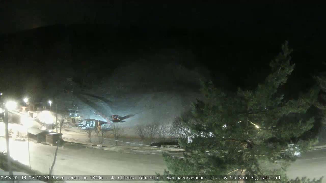 A nighttime, snow-covered ski slope is illuminated by artificial lights, with some small structures and a vehicle visible near the bottom.