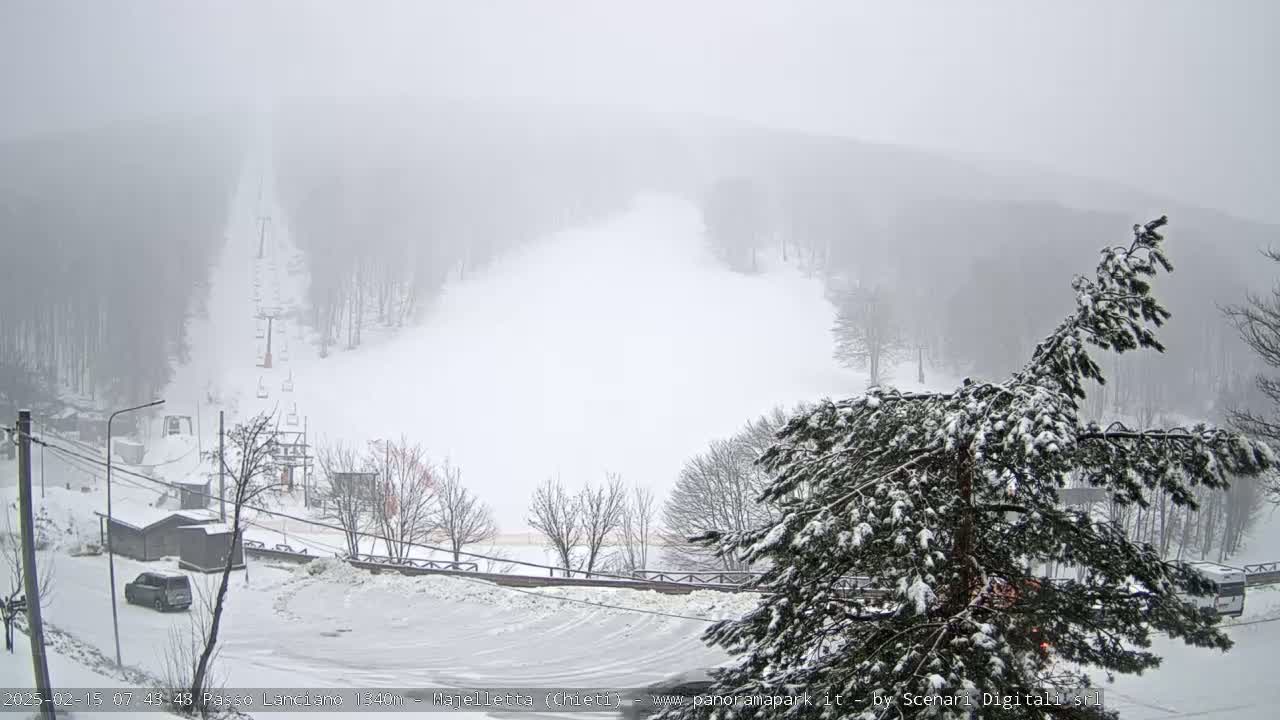 A snow-covered ski slope with a chairlift visible, surrounded by snow-laden trees in foggy conditions.