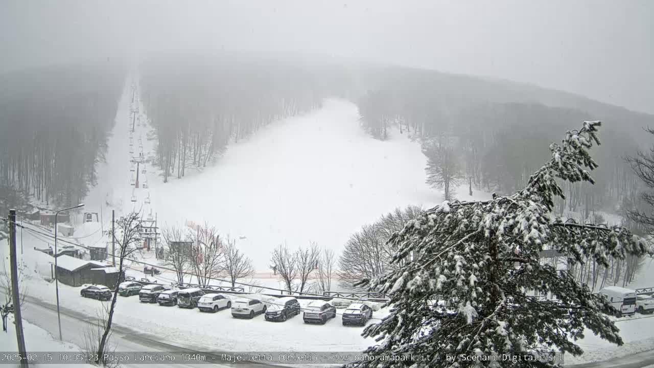 A snow-covered ski slope with a chairlift and a parking lot full of cars is visible through a light snowfall and fog.